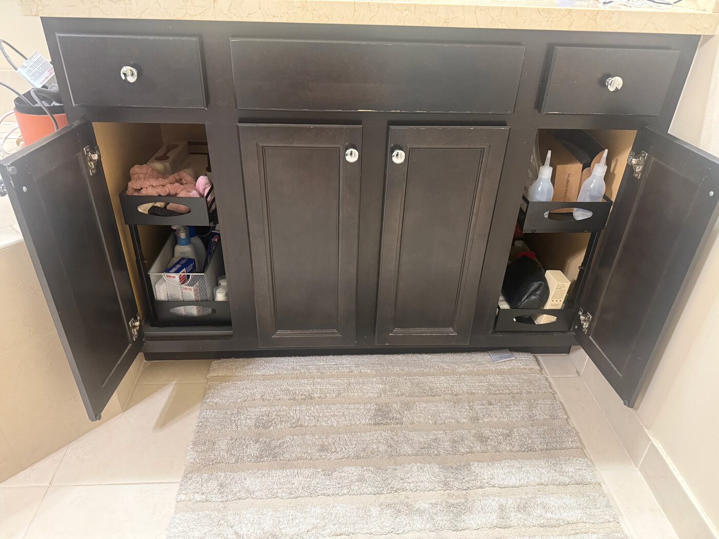 A double bathroom vanity with both outer doors open, showing one two-tier black sliding organizer in each side bay, away from the plumbing behind the closed center doors